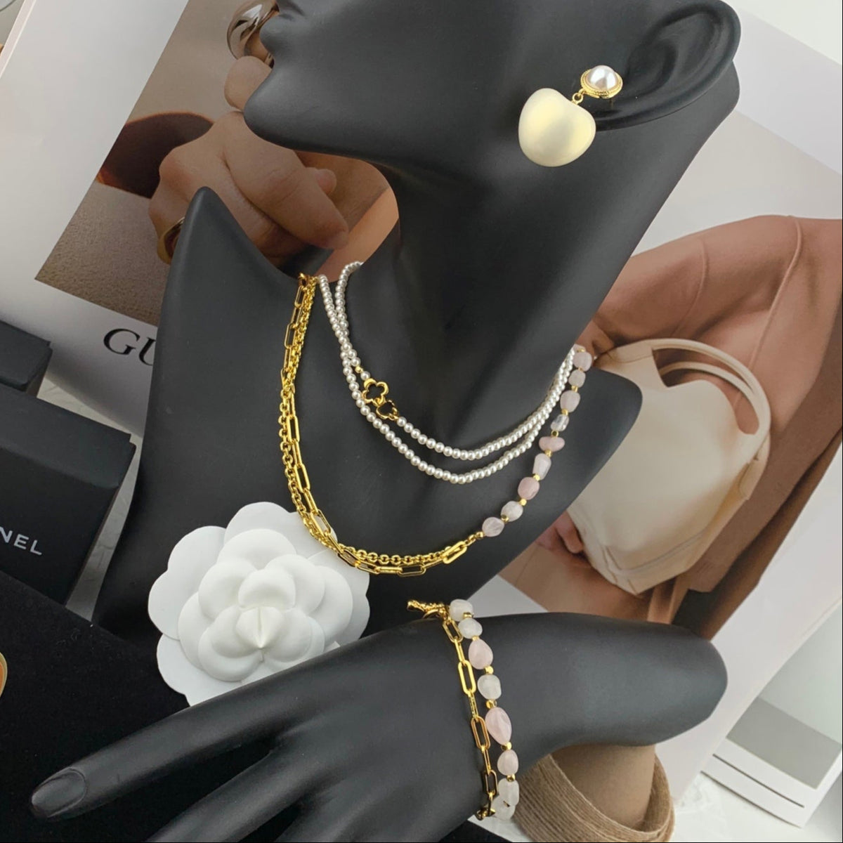Black mannequin hand with jewelry including a gold chain, pearl bracelet, and earrings on a blurred background.