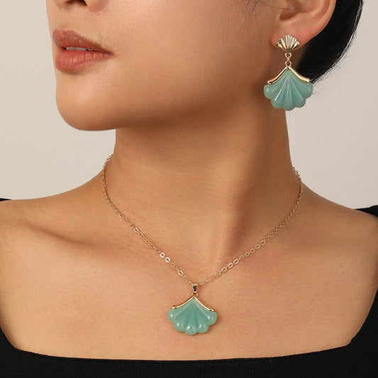 Close-up of a person wearing a gold necklace with a green pendant and green shell-shaped earrings.