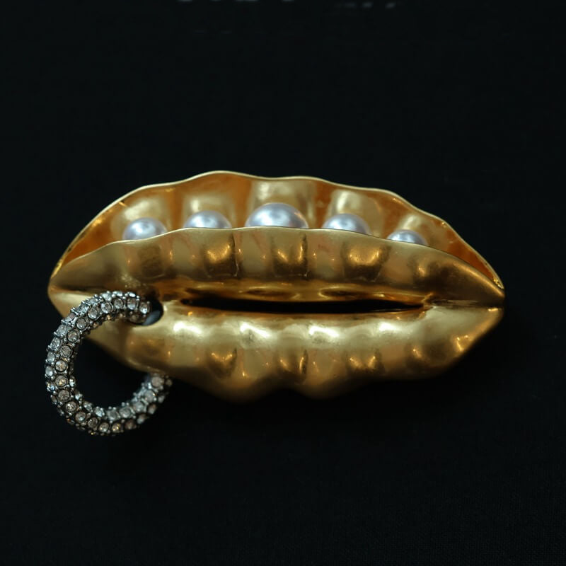 Gold lip-shaped brooch with pearls and a rhinestone ring on a black background