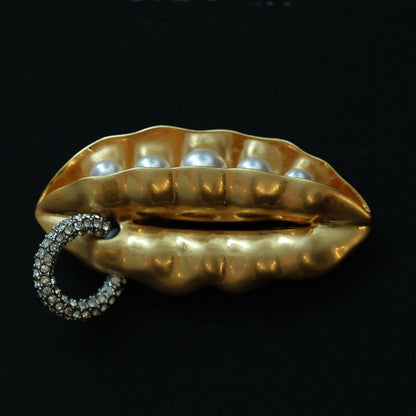 Gold lip-shaped brooch with pearls and a rhinestone ring on a black background