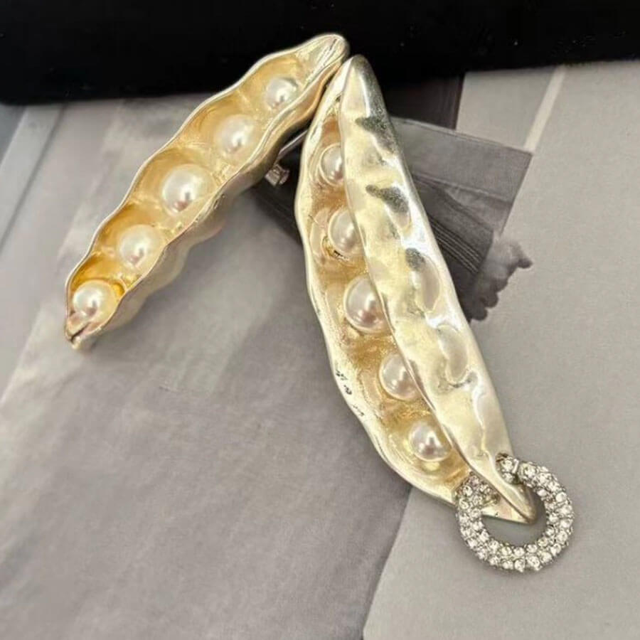 Gold earrings shaped like pea pods with pearl-like beads on a gray surface