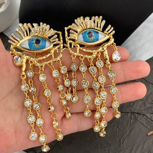 Gold eye-shaped earrings with blue gemstones held in a hand.